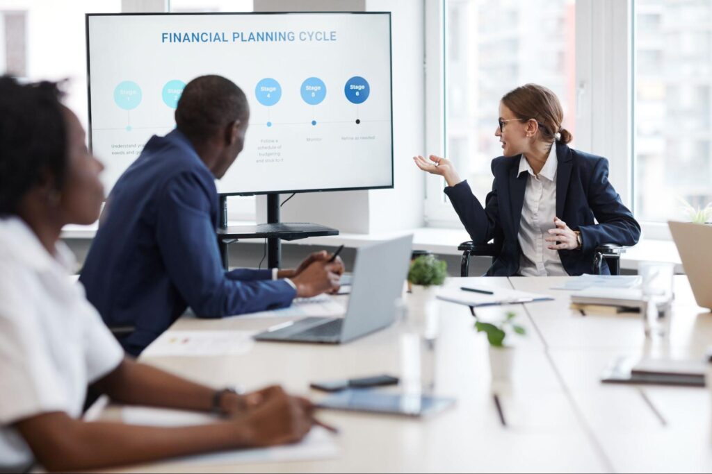 Corporate professionals in a conference room engaged in a presentation, with a presenter pointing to a large LCD screen.